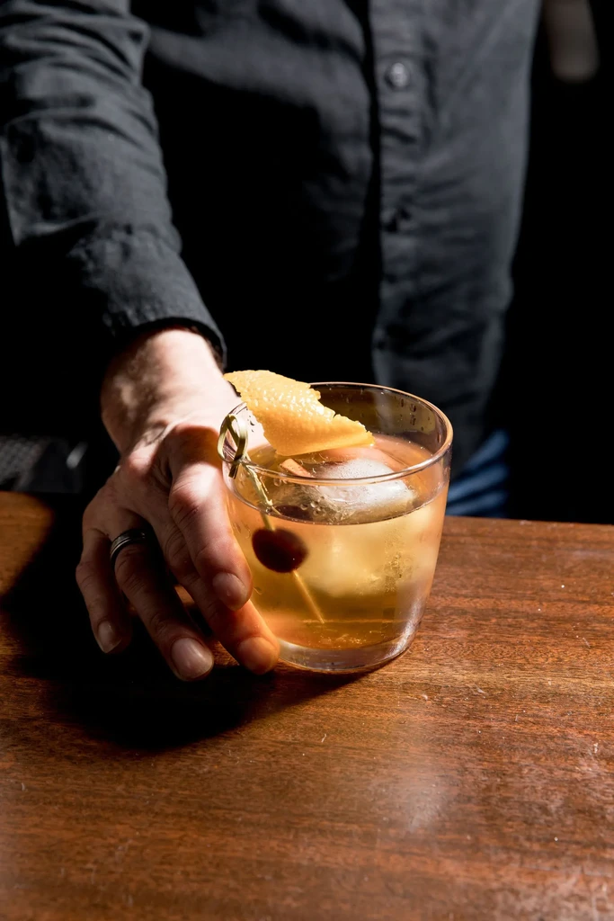 Close-up of amber cocktail with spherical ice, cherry, citrus peel.