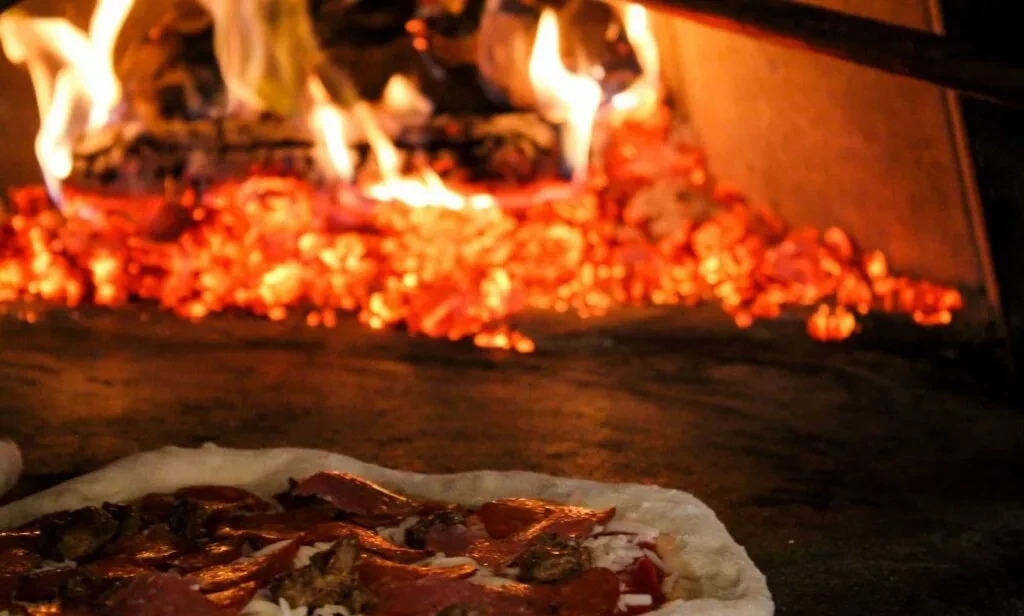 Uncooked pizza topped with meats, in a wood-fired oven with flames.