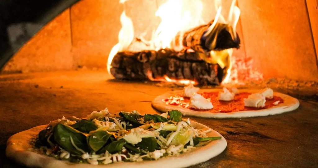 Raw spinach and tomato pizzas in a glowing wood-fired oven.