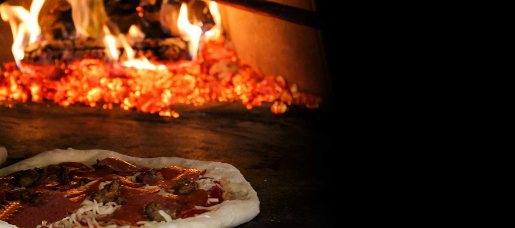 Uncooked pepperoni pizza on oven floor, illuminated by wood fire.