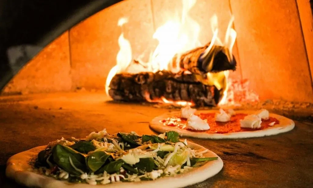 Two uncooked pizzas in a wood-fired oven, illuminated by flames.