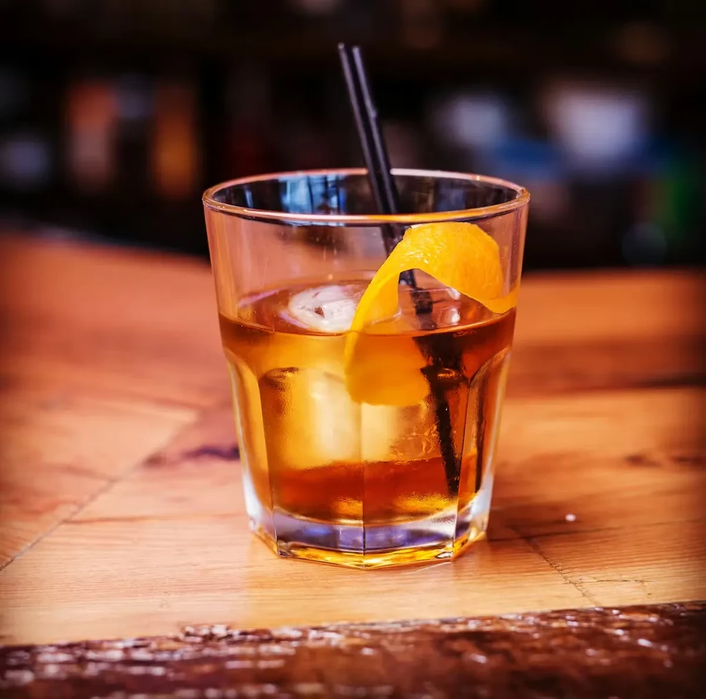 Amber cocktail in faceted glass with ice, orange peel, and straw.