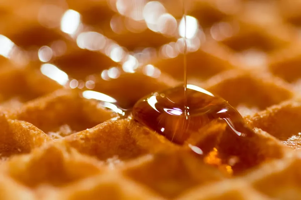 Close-up golden-brown waffle with amber syrup pouring, reflecting light.