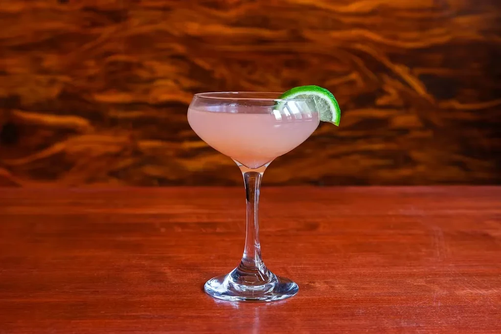 Pinkish-orange cocktail in coupe glass with lime on wooden table.