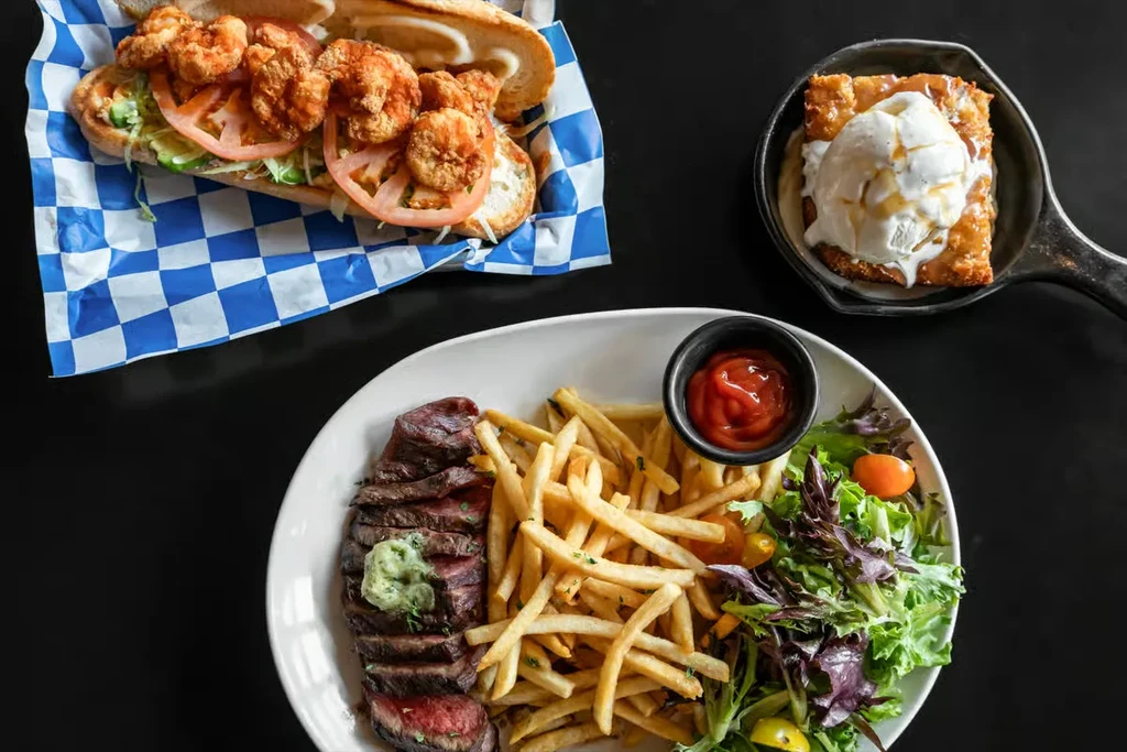 Overhead view of a shrimp po'boy, steak with fries, and skillet dessert.