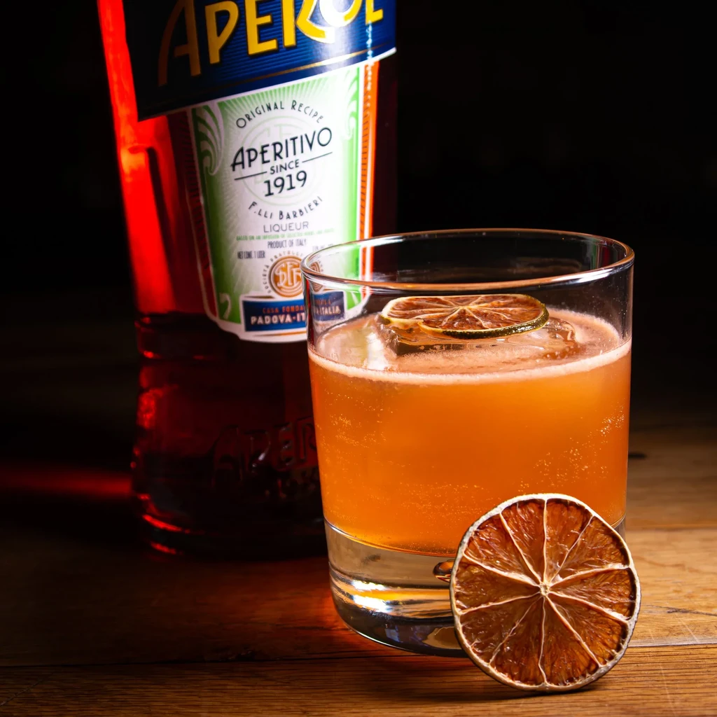Orange cocktail with dried citrus and Aperol bottle on wooden surface.