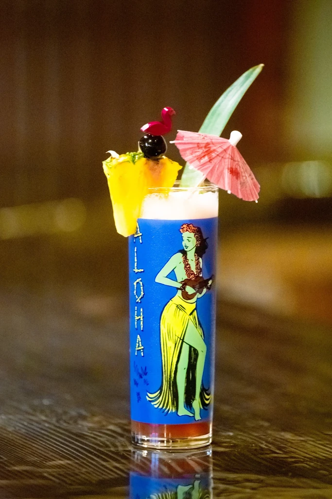 Layered blue cocktail in an ALOHA hula dancer glass, heavily garnished.
