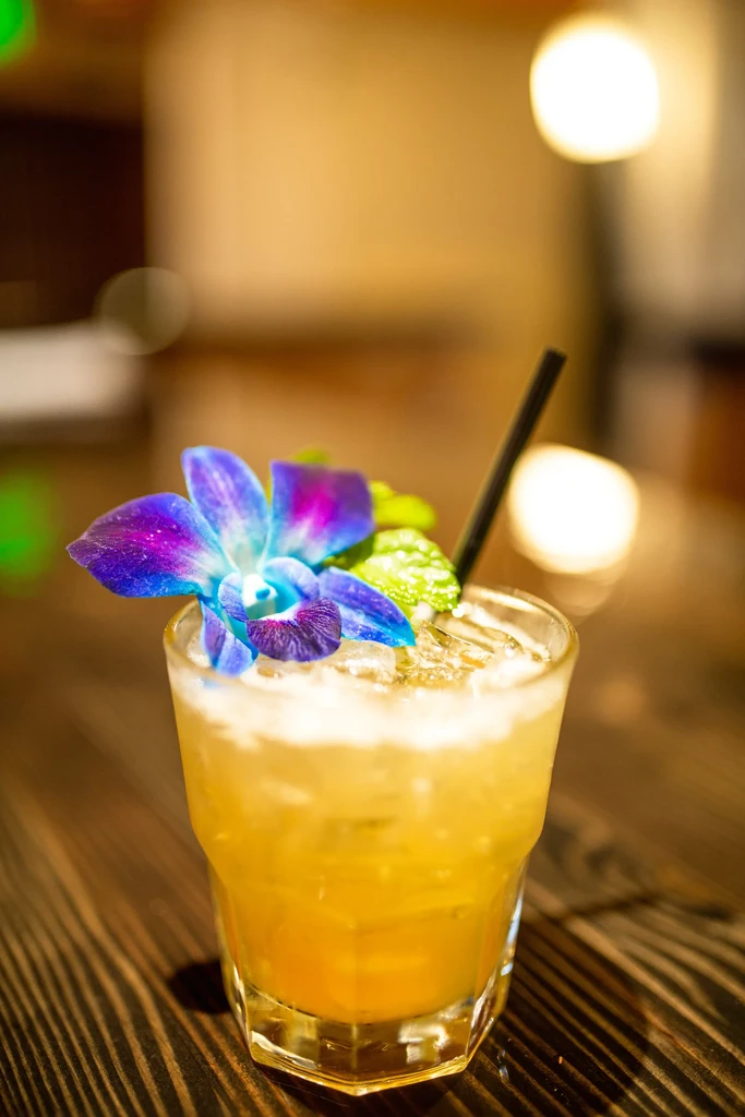 Frothy yellow-orange cocktail with a blue orchid and mint garnish.