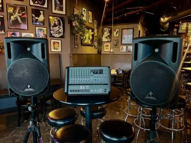 Dimly lit music bar with professional loudspeakers, mixer, and wall posters.