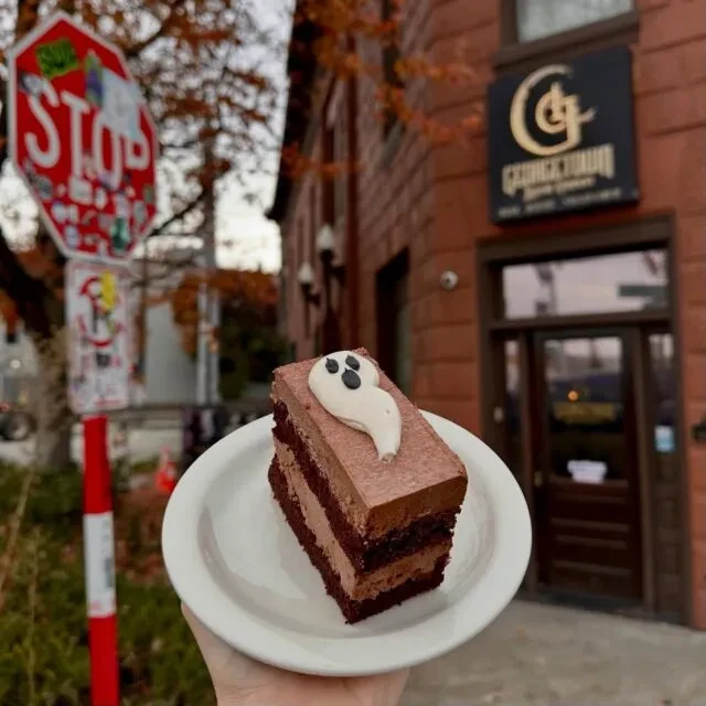 Hand holds a chocolate cake slice with a ghost decoration, autumn background.