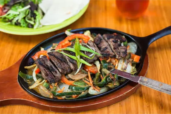 Sizzling steak fajitas in skillet, with salad, tortillas, and drink.