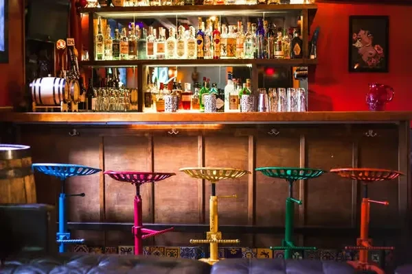 Brightly colored stools line a wooden bar filled with bottles and glassware.