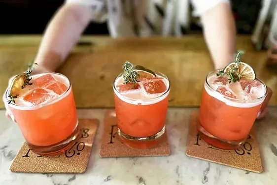 Row of three orange-red cocktails garnished with herbs and dried citrus.