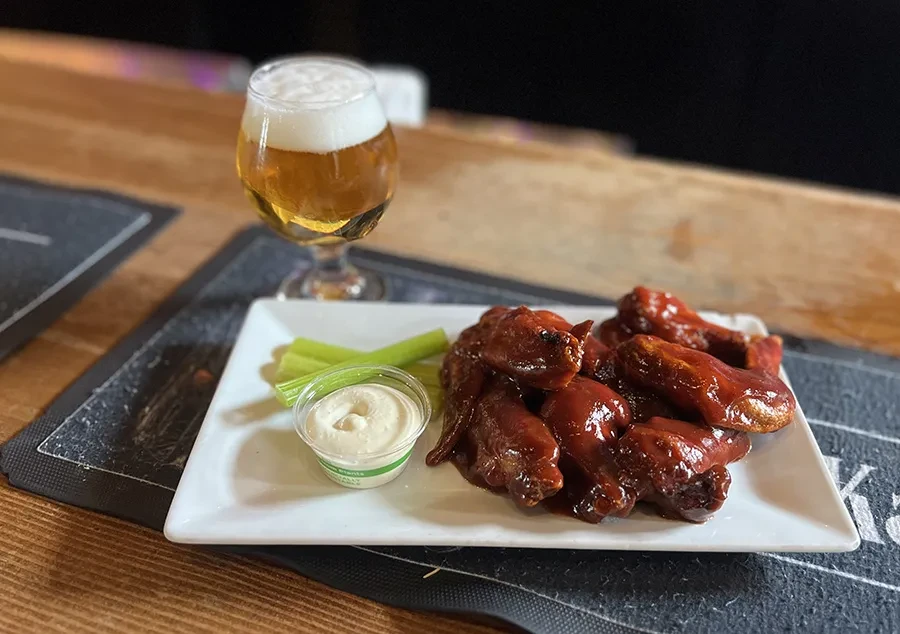 Plate of sauced chicken wings, celery, dressing, and beer on table.