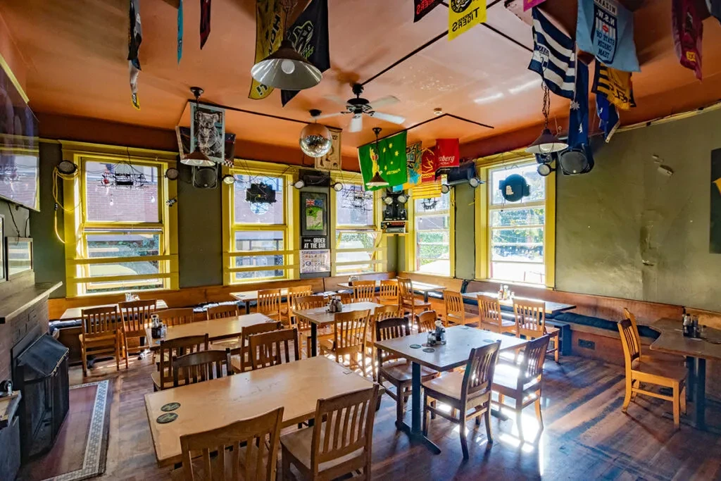 Rustic restaurant interior with wooden tables, olive walls, and bright yellow windows.