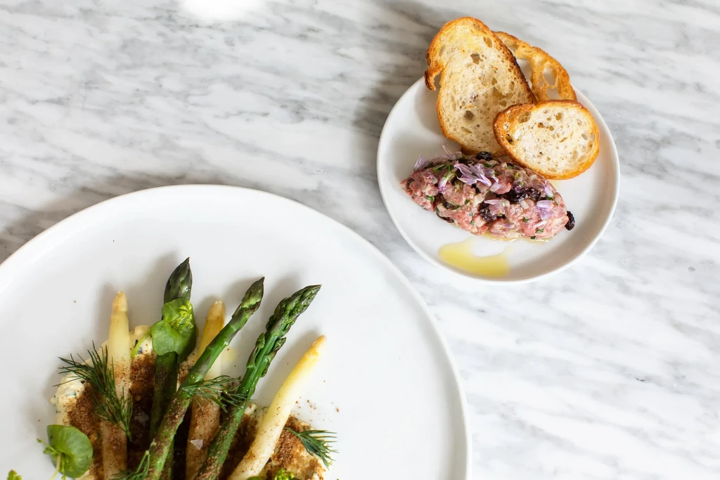 Overhead: Plated asparagus and tartare dishes on a white marble table.