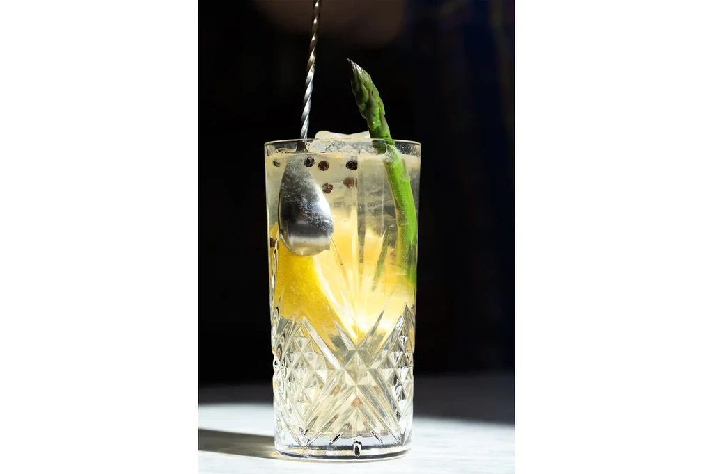 Cocktail in faceted glass, garnished with asparagus, lemon, and ice.