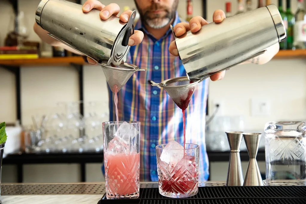 Bartender pours two colorful cocktails into ice-filled crystal glasses.