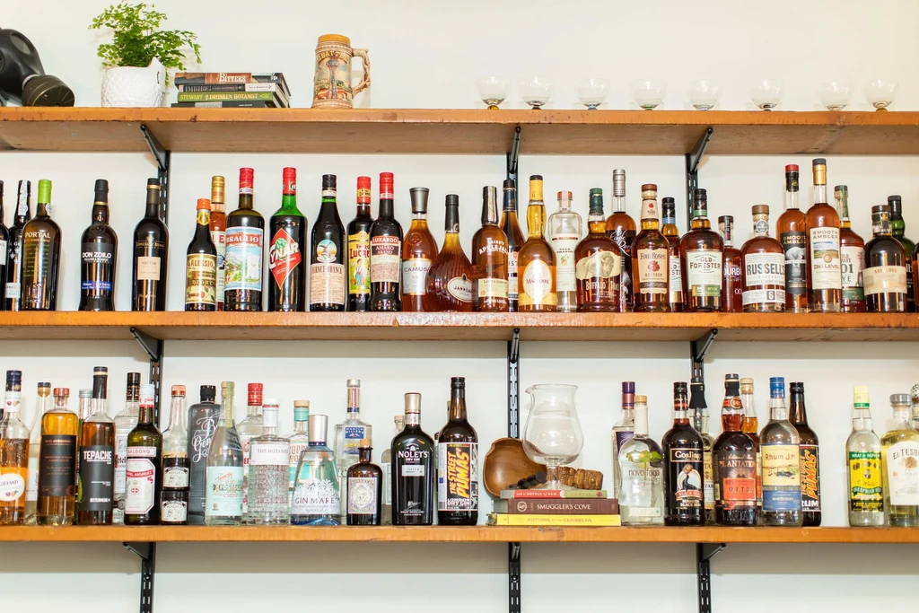 Well-stocked, tiered shelves display diverse liquor bottles, a gas mask, and books.