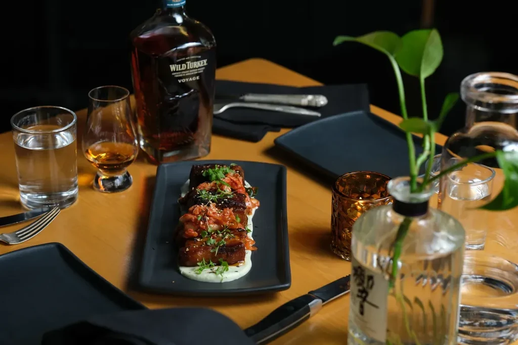 Braised meat dish, Wild Turkey bourbon, and whiskey on a warm restaurant table.