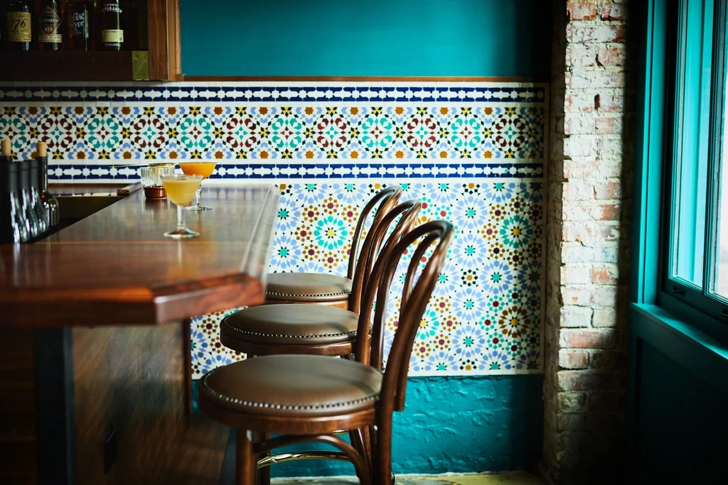 Polished wooden bar, two cocktails, leather stools, Moroccan tiled wall.