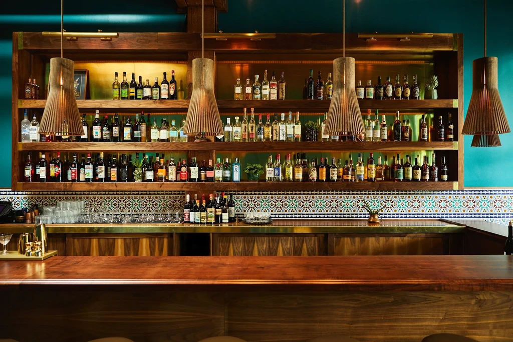 Restaurant bar area, bottles on wooden shelves, teal wall, geometric tiles.