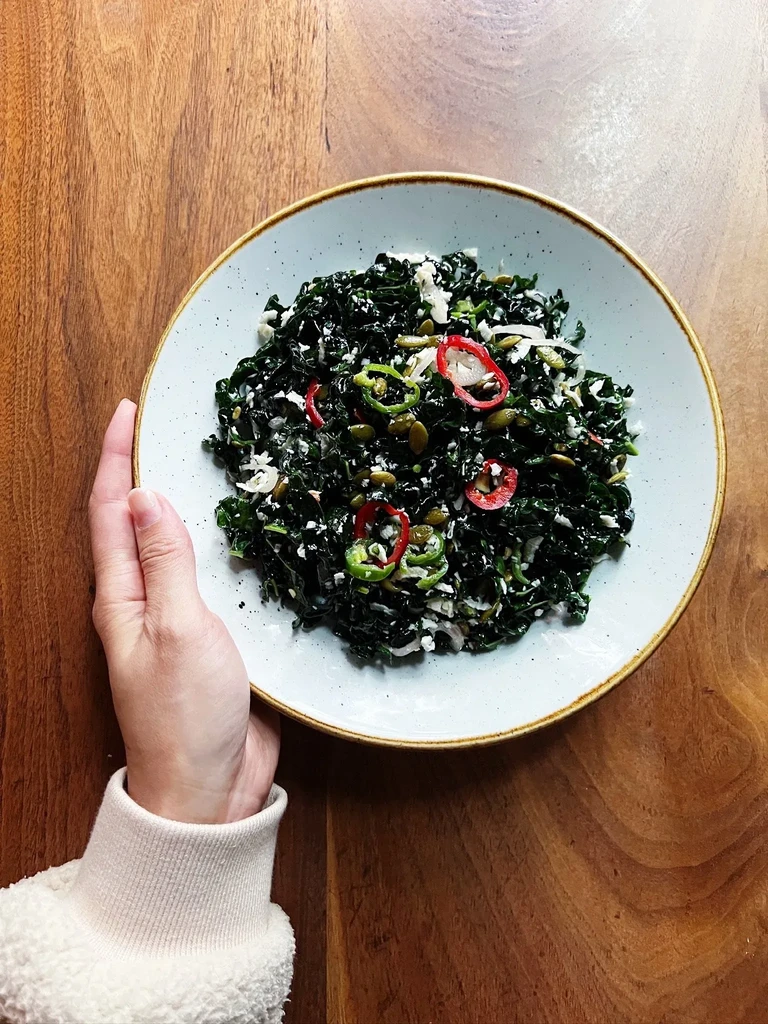 Hand holding bowl of kale salad with feta, peppers, and pumpkin seeds.