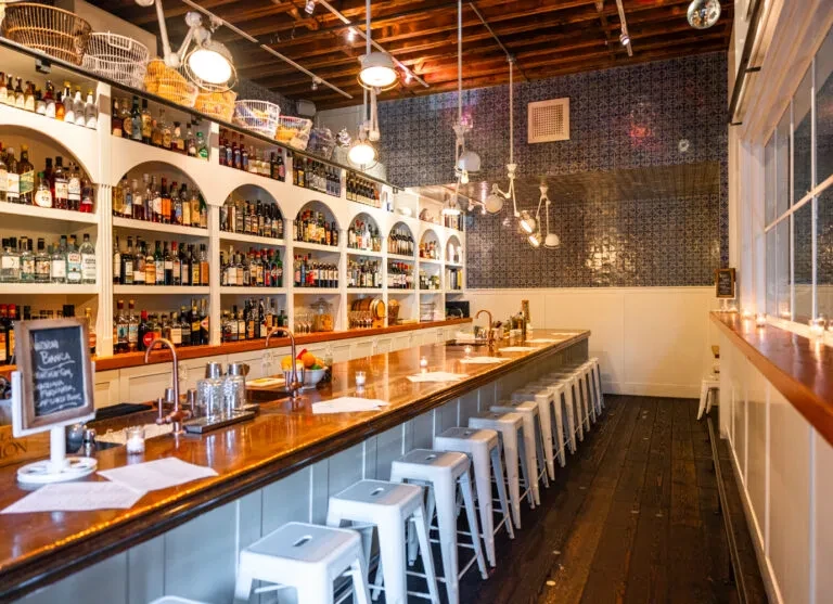 Stylish bar interior with wooden counter, white stools, and liquor bottles.