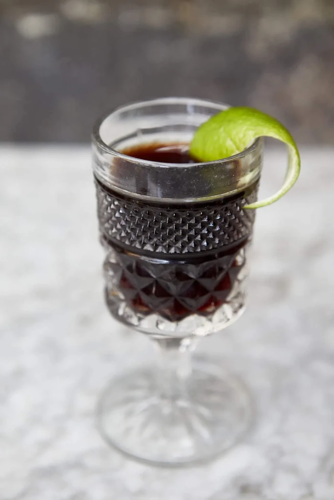 Dark cocktail in a faceted stemmed glass with a lime twist garnish.