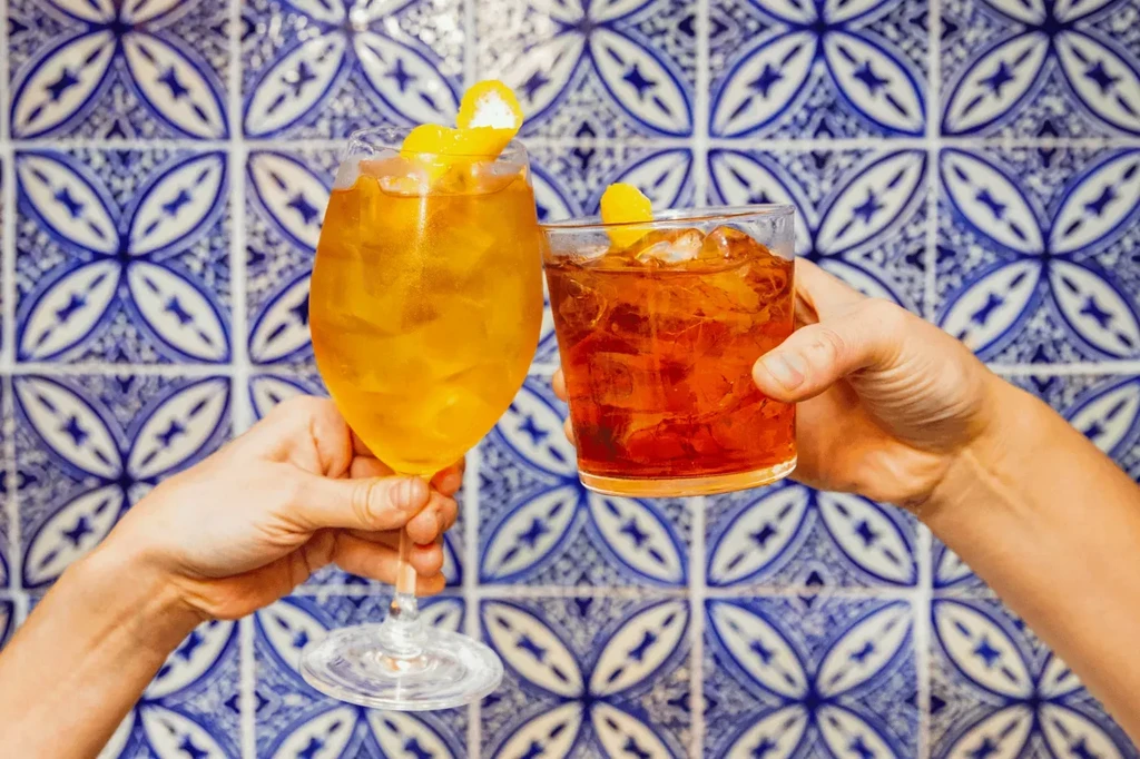 Hands toast two vibrant cocktails against patterned tiles.
