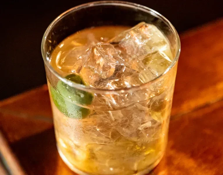 Amber drink with ice and lime wedge in a clear glass on wood.