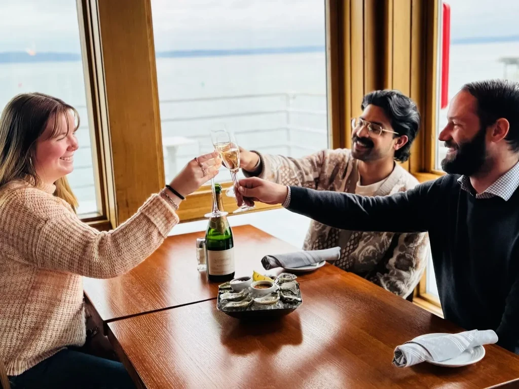 Three people toast with champagne and oysters at a waterfront restaurant.