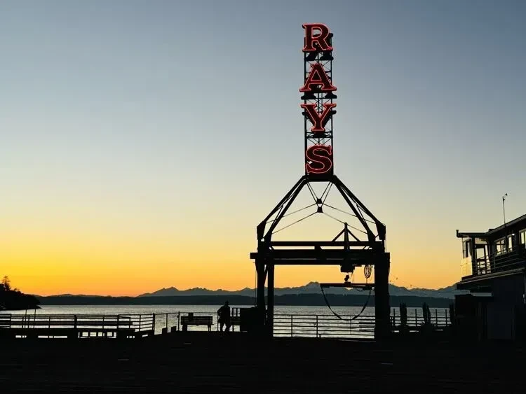 Neon 'RAYS' sign on a silhouetted pier with people at sunset.