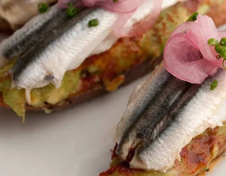 Two tapas with fish, green spread, pickled onions, and chives on bread.