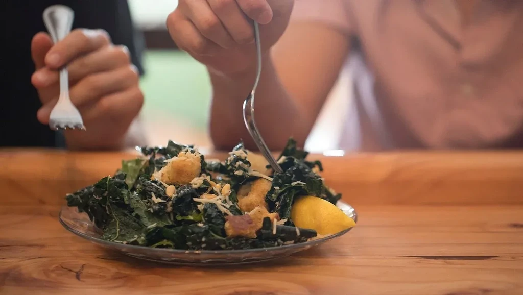 Hands eating a fresh kale Caesar salad with croutons on a wooden table.