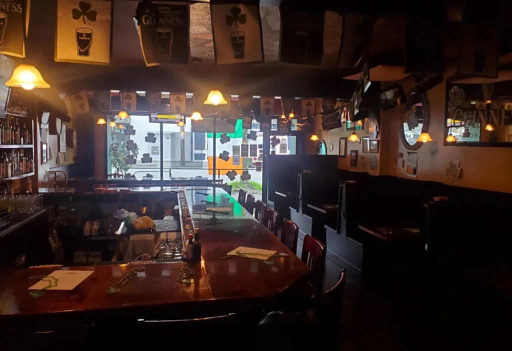 Dimly lit Irish pub interior with wooden bar, stools, and shamrock decor.