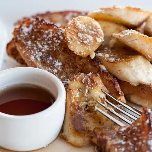 French toast topped with caramelized bananas, powdered sugar, syrup, and a fork.