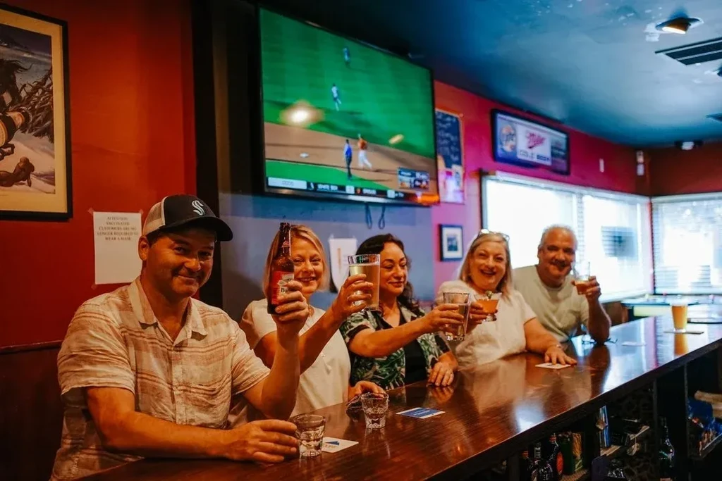 Five smiling adults toast drinks at a bar showing a baseball game.