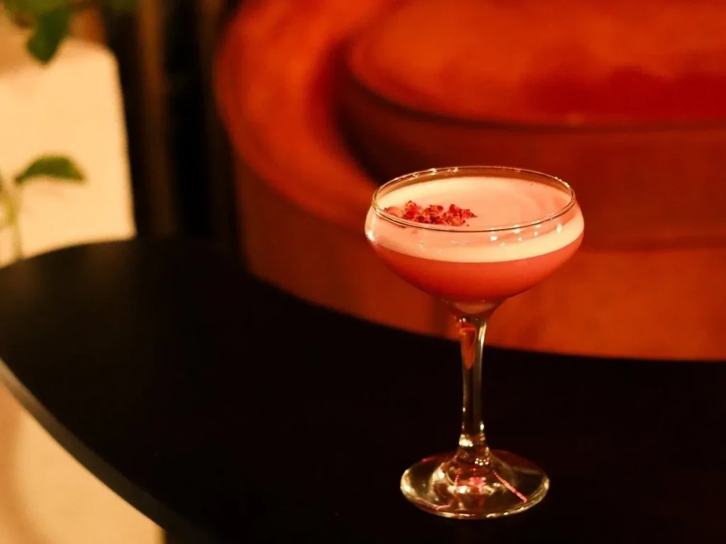 Pink cocktail in coupe glass, foamy top, rose petal garnish.