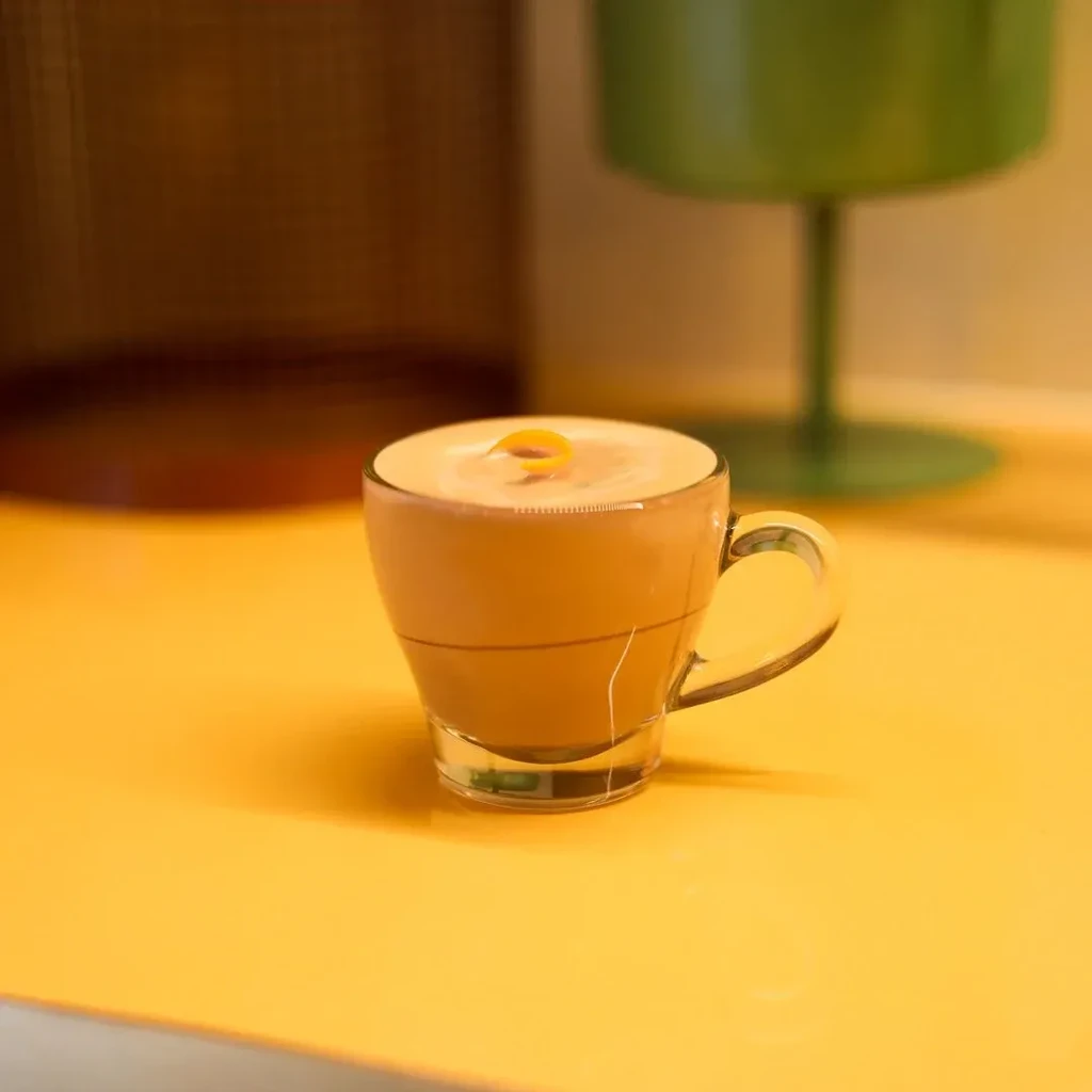 Clear glass mug with frothy drink and orange zest on yellow.