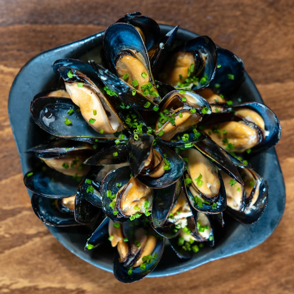 Cooked mussels garnished with chives in a dark bowl on a wooden table.
