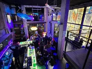 Multi-level industrial-chic bar, vibrant neon lights, people, and art installation.