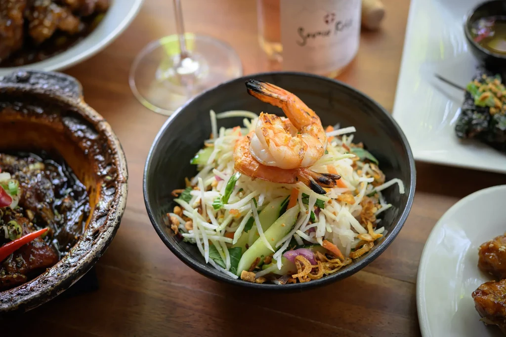 Shrimp salad, dark saucy dish, and rosé wine on a wooden table.