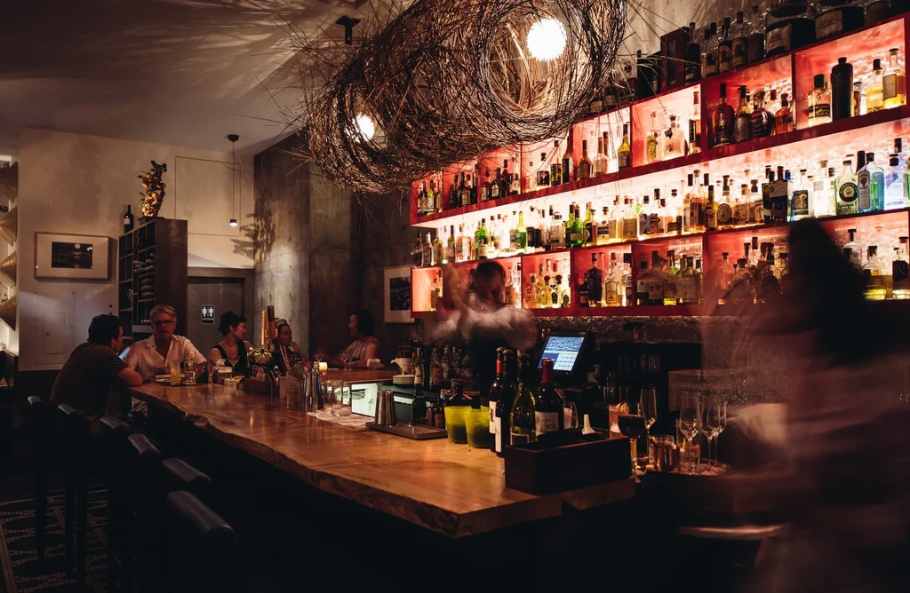 Intimate bar with blurred bartender shaking cocktail, red-lit liquor.