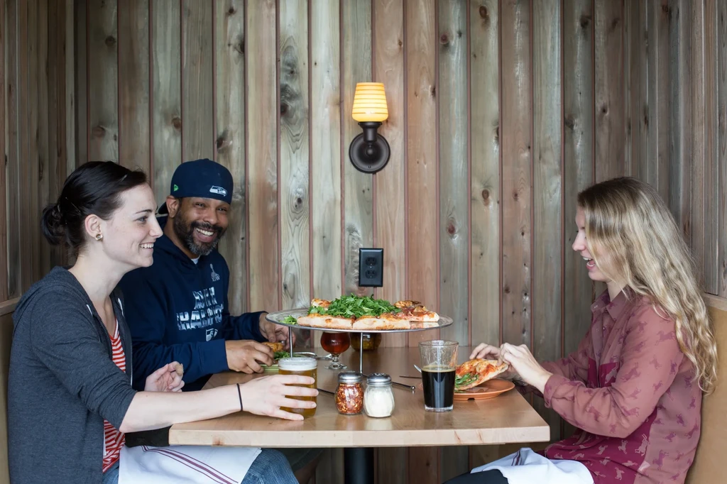 Three smiling people with pizza and drinks in a restaurant booth.