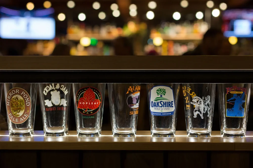 Empty pint glasses with distinct brewery logos on a lit wooden bar.