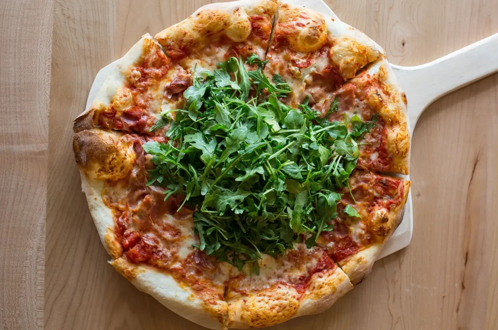 Sliced pizza with arugula, cured meat, and cheese on wooden peel.