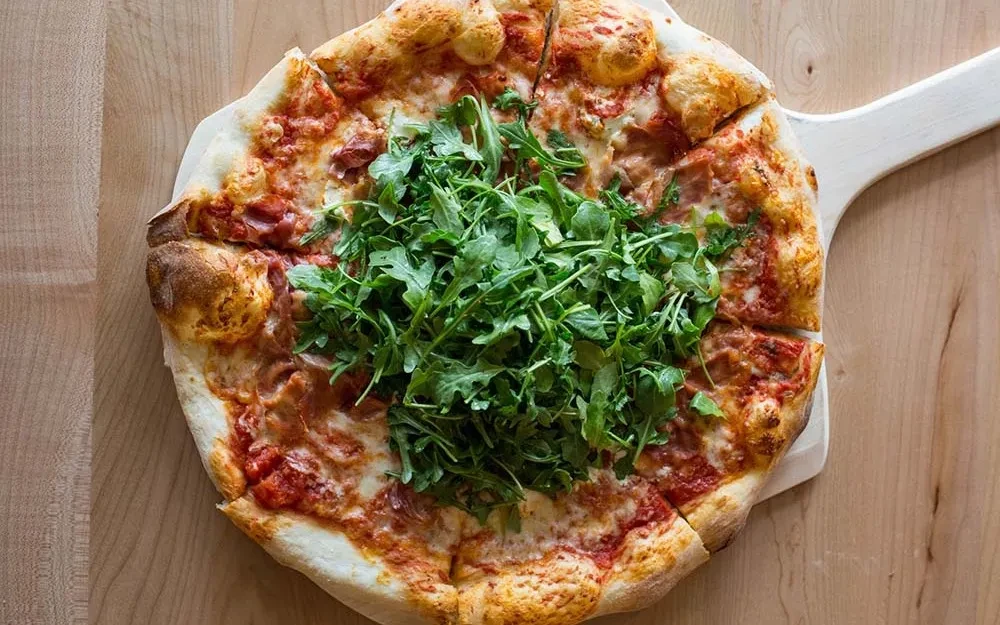 Sliced pizza with tomato, cheese, arugula, and meat on a peel.