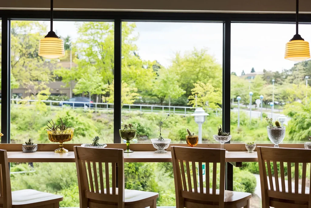 Restaurant seating area with plants, wooden counter, and green outdoor view.