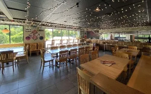 Empty restaurant dining area with wooden tables, chairs, windows, and string lights.